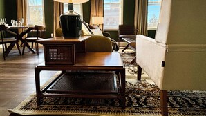 Un salon chaleureux avec une table basse en bois, une lampe et un canapé. Il y a deux fenêtres avec des rideaux, une vue sur un immeuble à l’extérieur et un tapis à motifs sur le sol.