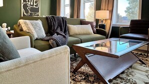 Un salon avec un mur vert, un tapis à motifs, une table basse en verre et un canapé agrémenté de plusieurs coussins.