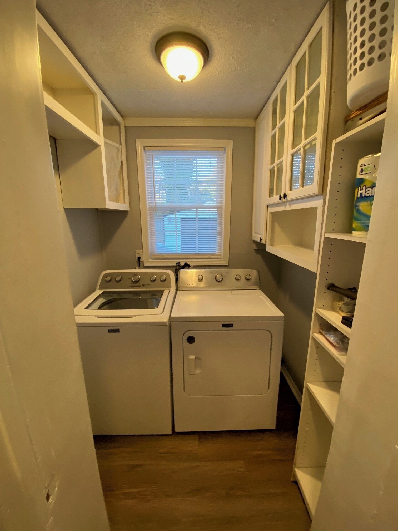A laundry room with a washer and dryer, a window with blinds, and shelving with laundry supplies.