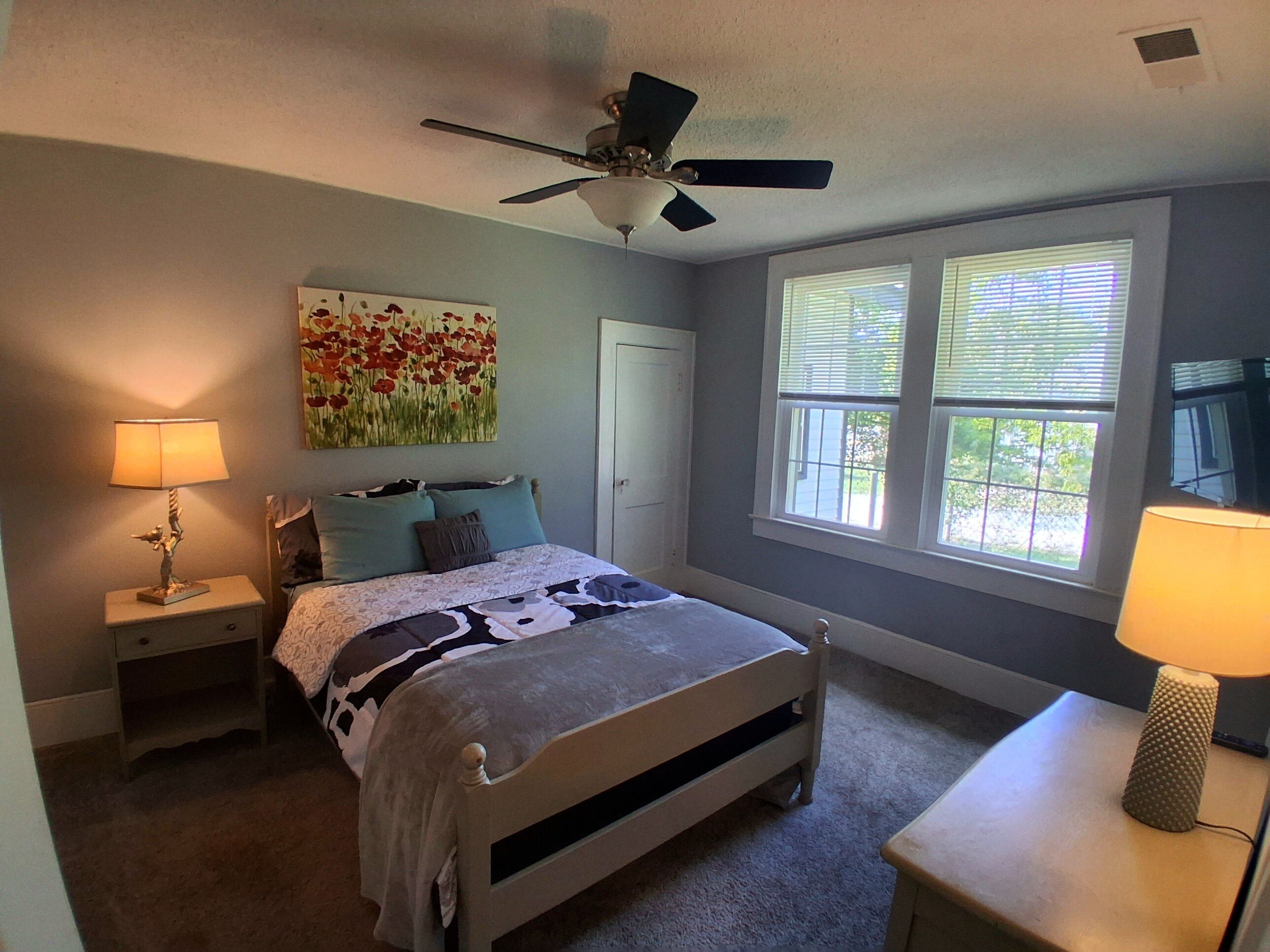 A bedroom with a bed, a nightstand with a lamp, a ceiling fan, a window with blinds, and a door.