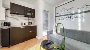 A modern kitchen with dark cabinets, a microwave, and a coffee maker. There is a grey sofa with a decorative wall art piece and a potted orchid. - TBS 76 - Superb studio in the Marais district (Paris)