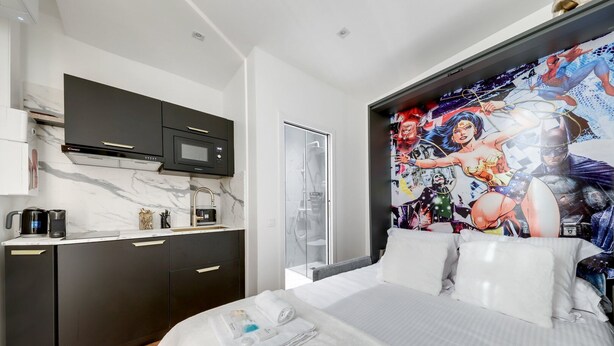 A modern hotel room with a bed, a black minibar, a microwave, and a large poster of comic book characters on the wall. - TBS 76 - Superb studio in the Marais district (Paris)
