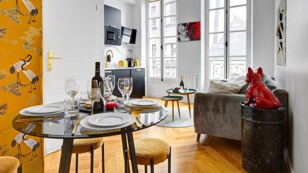 Un comedor con una mesa de vidrio redonda dispuesta para seis comensales, una cocina al fondo y una figurita de perro rojo sobre una mesita auxiliar.