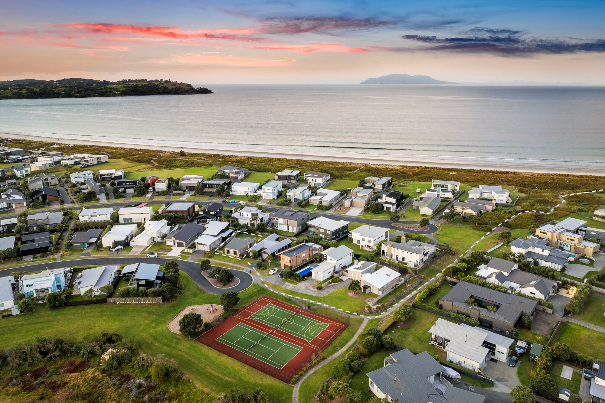 An aerial view of a coastal residential area with modern houses, a tennis court, and a beach in the background.