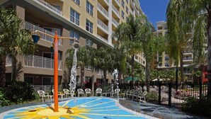 A swimming pool with a sun design, surrounded by lounge chairs and palm trees, with a hotel building in the background. - Vacation Village Reservations (Kissimmee)