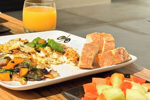 Een ontbijt bord met roerei, gebakken groenten en toast, vergezeld van een glas sinaasappelsap en een portie blokjes fruit.