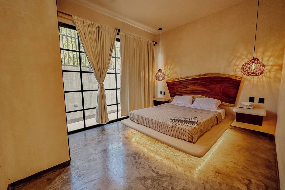 A bedroom with a large bed, a nightstand, a bedside table, and a window with curtains.