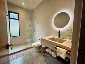 A modern bathroom with a glass-enclosed shower, a white toilet, a beige sink with a round mirror, and a wooden shelf with towels. - Casa Kobaab Tulum (Tulum)