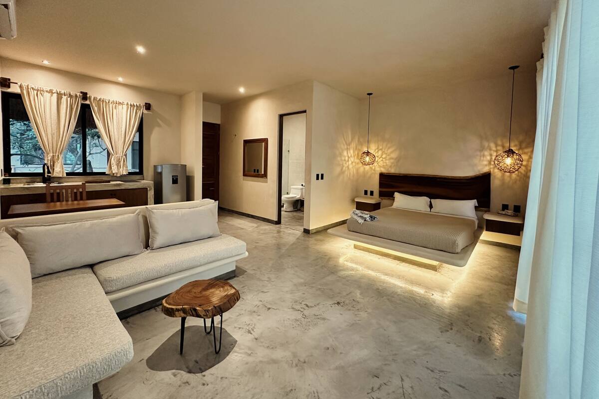 A modern hotel room with a sofa, a wooden coffee table, a bed with a headboard, and pendant lights.