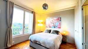 Une chambre à coucher avec un lit, une lampe de chevet, un luminaire fixé au mur, une fenêtre avec des rideaux et une vue sur le balcon.