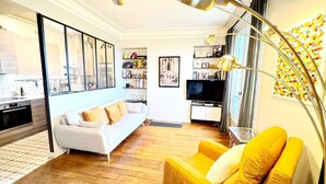 Un salon moderne avec un canapé, un fauteuil jaune, un téléviseur et une bibliothèque.