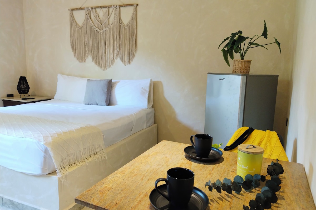 A neatly arranged hotel room with a bed, a small table with a mug and a plant, and a macrame wall hanging.