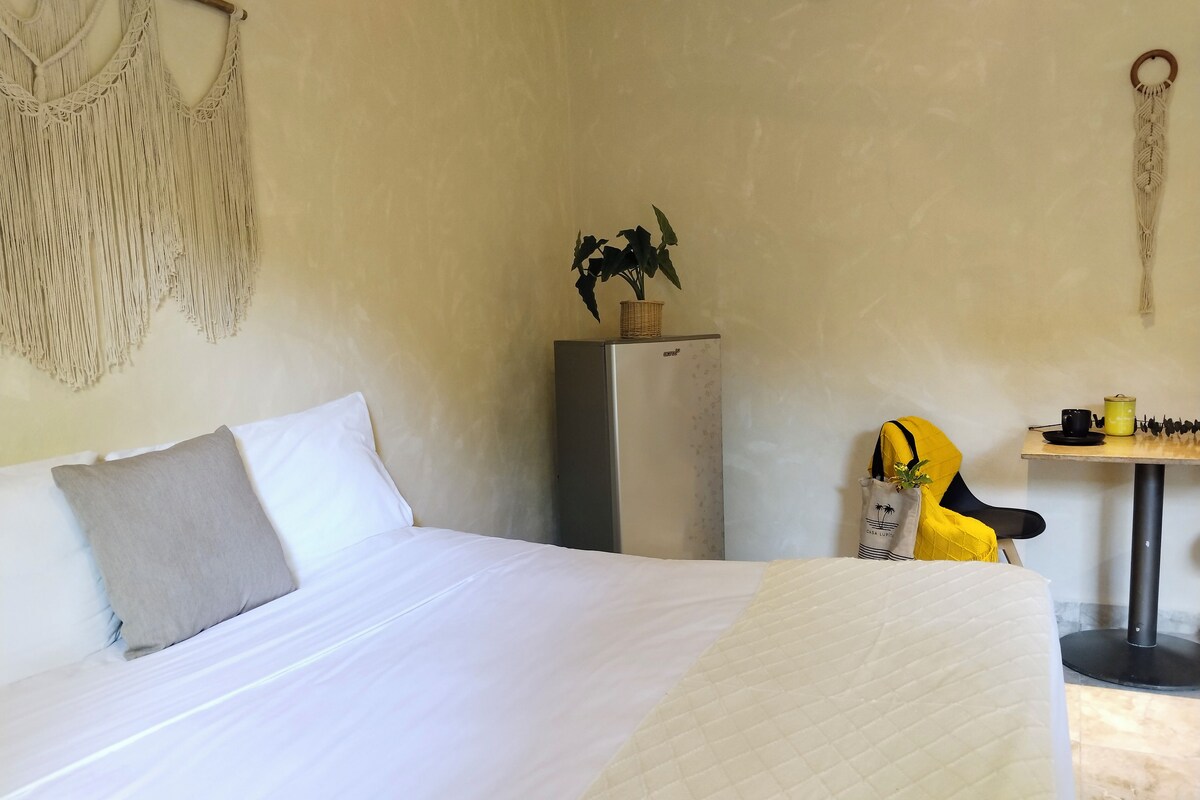 A hotel room with a bed, a small table with a yellow chair, a refrigerator, and a macrame wall hanging.