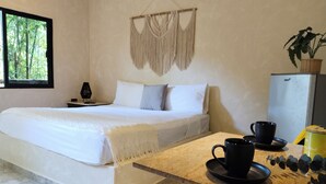 Un dormitorio con cama, mesita de noche, lámpara, colgador de macramé en la pared y vista a plantas a través de una ventana.