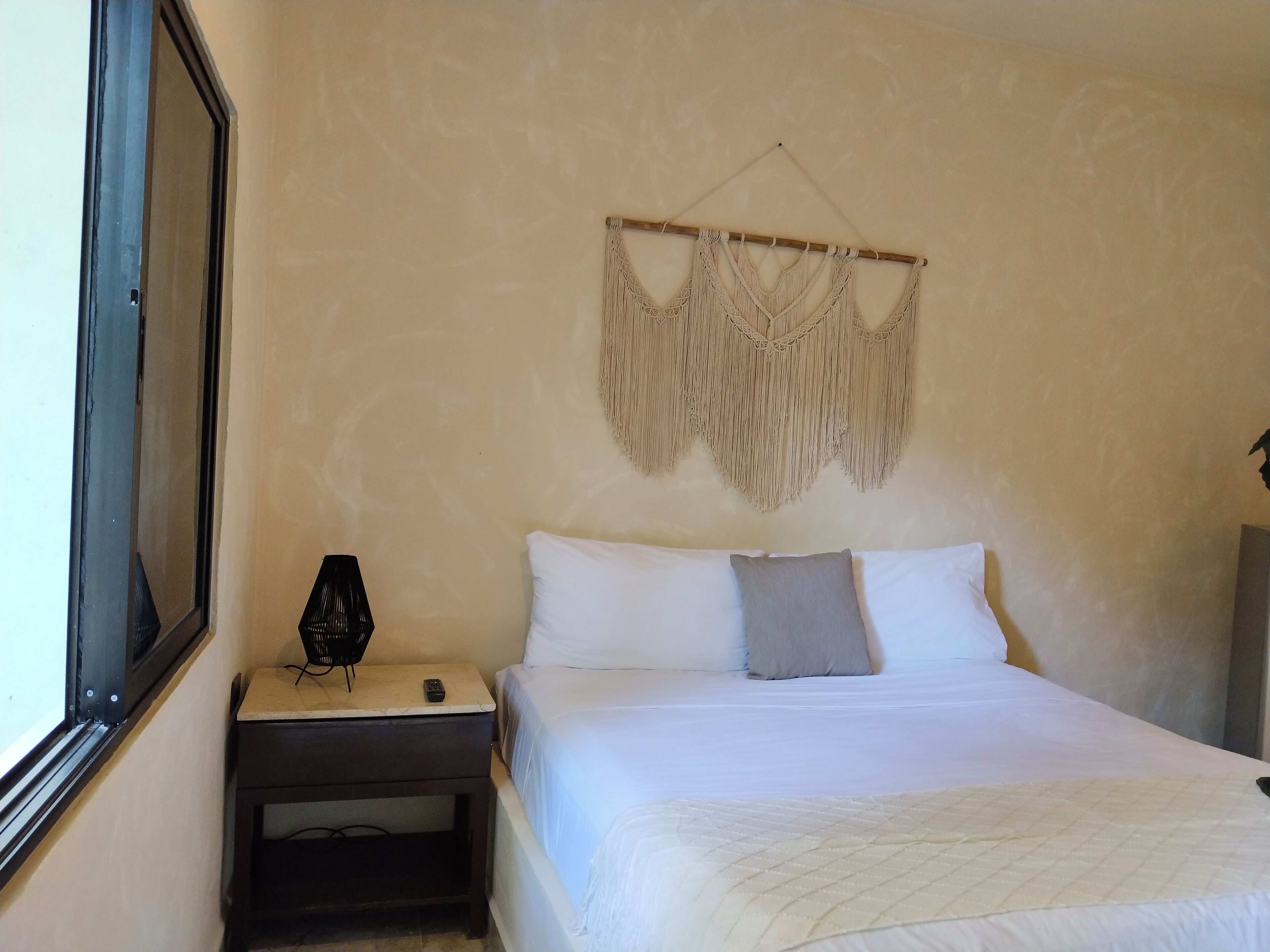 Un dormitorio con cama, mesita de noche, un adorno de macramé en la pared y una ventana.