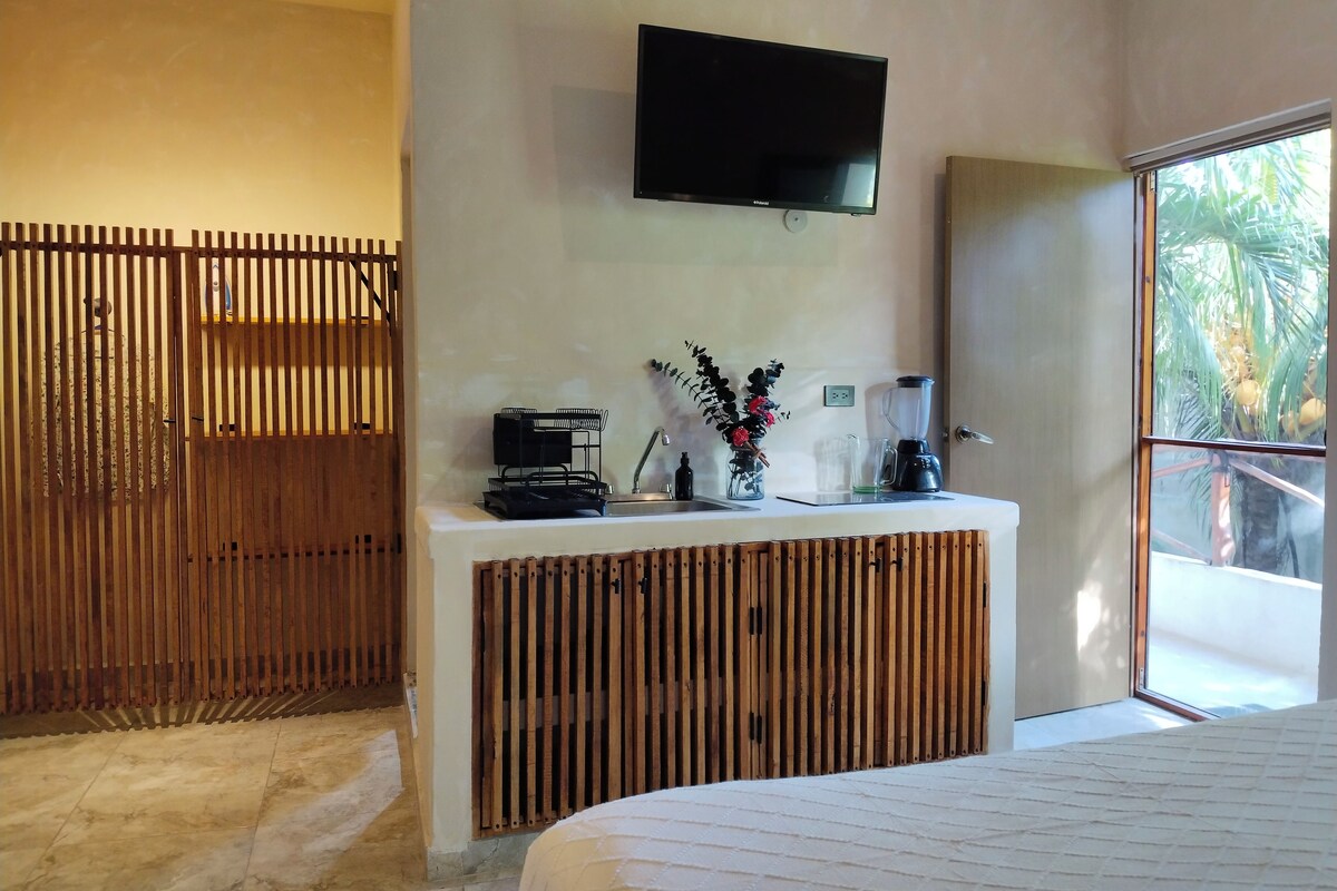 A modern hotel room with a large bed, a flat-screen TV mounted on the wall, a wooden partition, and a small kitchenette area.