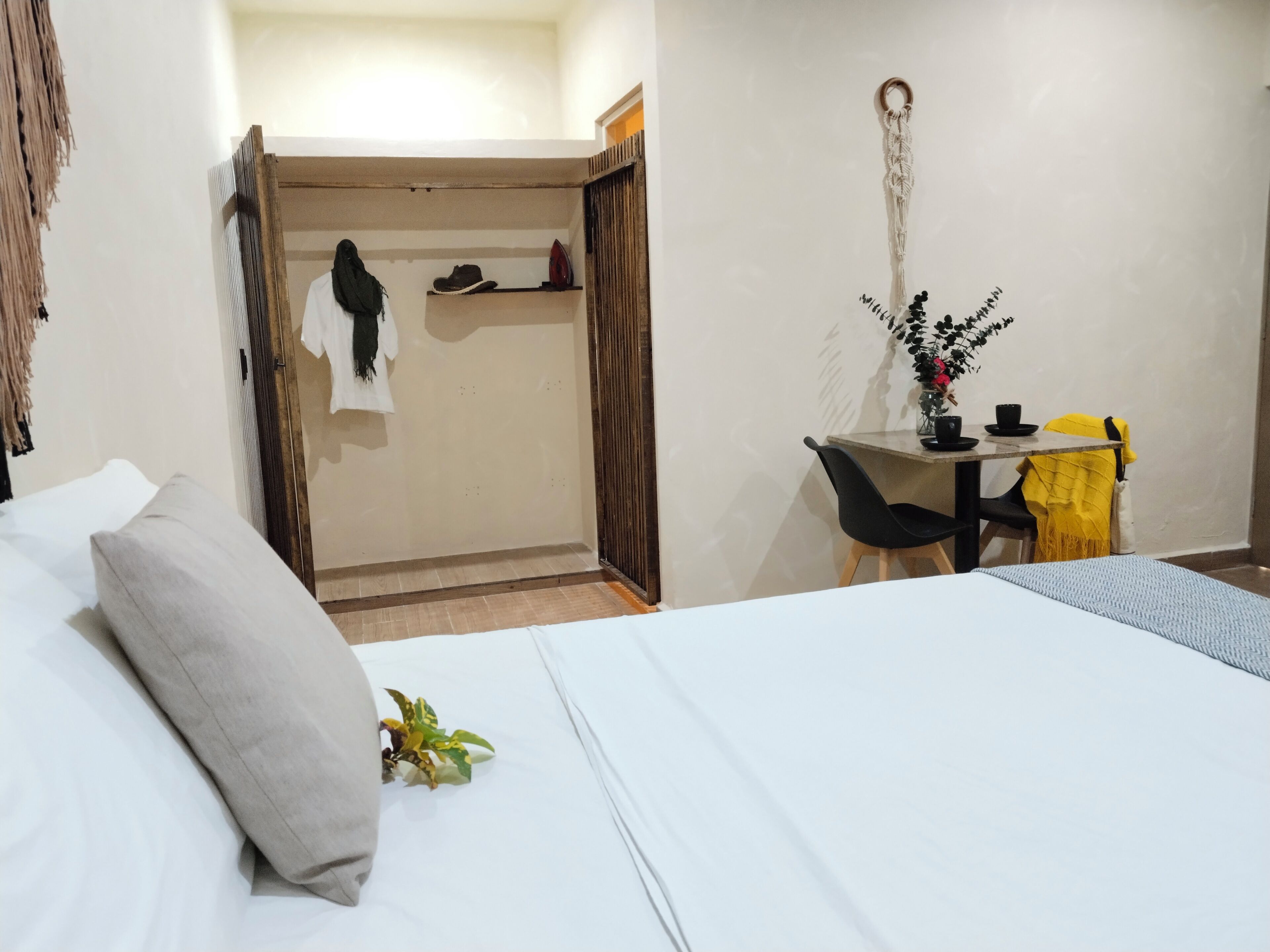 Habitación de hotel con cama, escritorio y una silla. Hay una planta sobre la cama y una decoración de macramé en la pared.