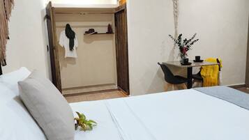 Habitación de hotel con cama, escritorio y una silla. Hay una planta sobre la cama y una decoración de macramé en la pared.