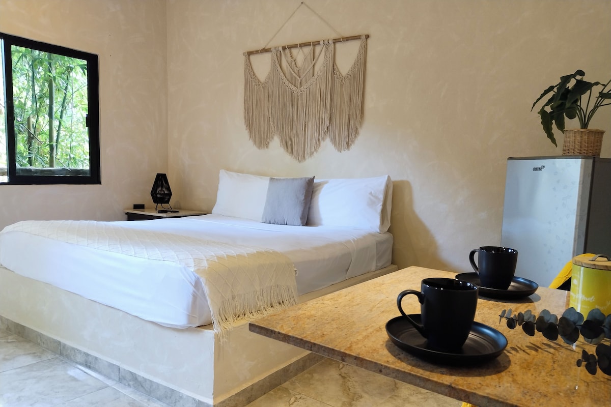 A bedroom with a bed, bedside table, lamp, macrame wall hanging, and a view of greenery through a window.