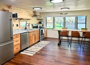 A modern kitchen with wooden cabinets, stainless steel appliances, and a large window providing a view of the outdoors.