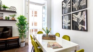 A modern dining area with a white table, green chairs, and a magazine on the table. There are potted plants and a TV on a shelf. - 3 Bd Apartment With Balconies Next to Larios Street Larios (Málaga)