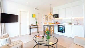 A modern living room with a glass coffee table, a sofa, and a TV. A kitchen with white cabinets and appliances is visible in the background. - 2 Bedroom With Private Terrace and Communal Pool in Marbella Center Ricardo Soriano Terrace (Marbella)