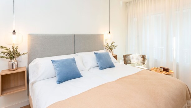 A neatly made bed with a grey headboard, white and blue pillows, and a beige blanket. There are bedside tables with lamps and a small plant on one side. - Amazing Apartment 5 Bd in the Heart of Granada Alh Ndiga (Granada)