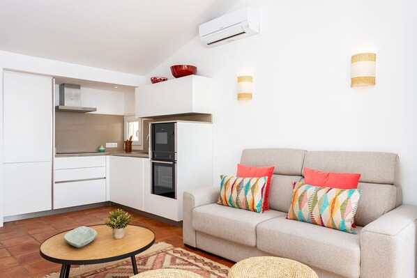 Eine moderne Küche mit weißen Schränken, ein graues Sofa mit bunten Kissen und ein kleiner runder Tisch mit einer Pflanze.