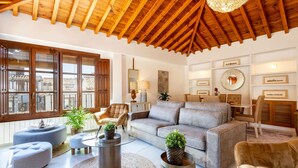 Ein geräumiges Wohnzimmer mit hoher Holzbalkendecke, einem großen grauen Sofa, zwei Sesseln und einem kleinen Esstisch. An den Wänden befinden sich Kübelpflanzen und ein runder Spiegel.