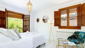 Ein Schlafzimmer mit Bett, Nachttisch, Lampe, Stuhl und großen Fenstern mit Holzläden.