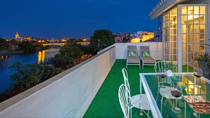 Eine Dachterrasse mit einem Glastisch, gedeckt für ein Abendessen, mit Blick auf einen Fluss und eine Stadtlandschaft im Abenddämmerung.