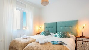 Een tweepersoonsbed met een lichtblauwe hoofdbord, twee kussens, een nachtkastje met een lamp en een raam met witte gordijnen.