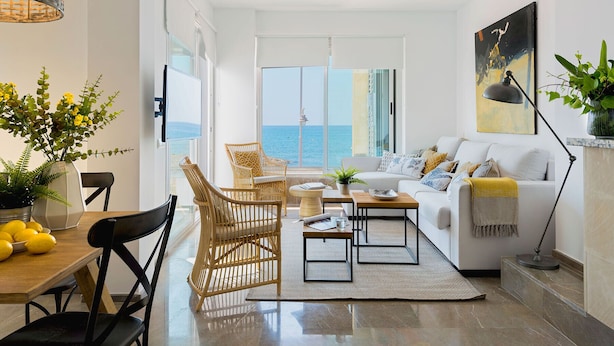 Una sala moderna con un sofá blanco, sillas de mimbre, una mesa de comedor con limones y vista al mar.