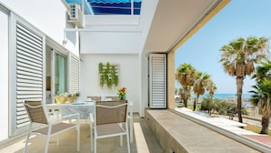 Un balcón al aire libre con una mesa y sillas, vista a la playa y palmeras.