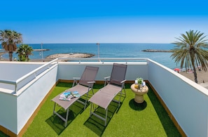 Ein Balkon mit zwei Stühlen und einem Tisch, der auf einen Strand mit Palmen und wolkenlosen, blauen Himmel blickt.
