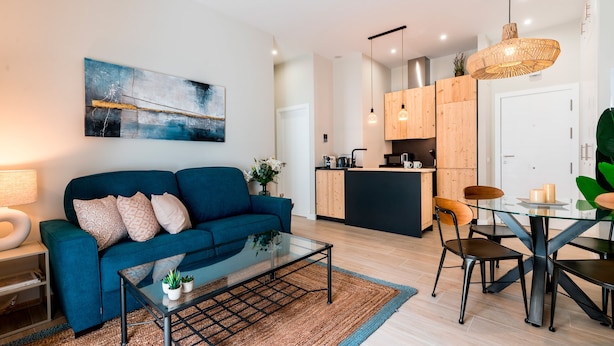 A modern living room with a blue sofa, a glass coffee table, and a dining area with a glass table and chairs. - Beautiful 1 bd Apartment in the Historic Center of Cadiz Sagasta (Cádiz)
