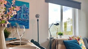 En matplats med ett bord dukat för fyra personer, en målning som föreställer en stadsbild, en lampa och en utsikt över grönskan genom ett fönster.