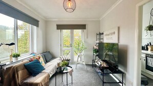 Ett mysigt vardagsrum med en soffa, ett soffbord och en tv. Det finns ett fönster med utsikt över träd och en bokhylla med böcker och dekorativa föremål.
