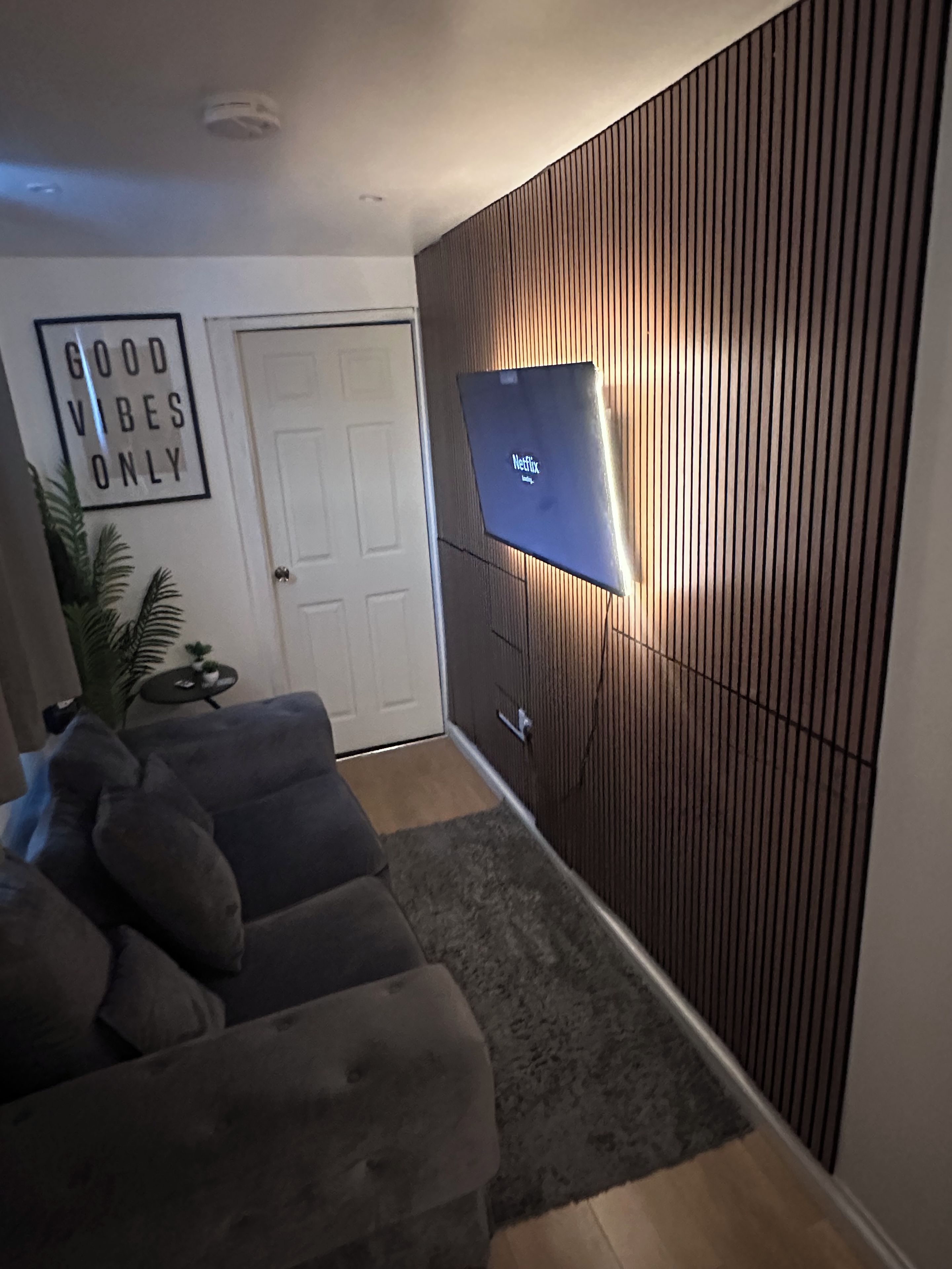 A living room with a grey sofa, a wall-mounted TV displaying the Netflix logo, a door, and a wall with vertical wooden slats.