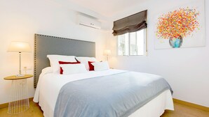 Un lit bien fait, avec des oreillers blancs et rouges, une tête de lit grise et un plaid bleu. On y trouve une table d’appoint ronde avec une lampe, une fenêtre agrémentée d’une lambrequin marron et un climatiseur fixé au mur.