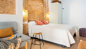 A cozy bedroom with a brick wall, a bed with white bedding, a grey sofa, and a small stool. - Cozy Studio in Santa Cruz Quarter Santa Teresa I (Seville)