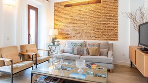Una sala de estar con pared de ladrillo, un sofá, dos sillones, una mesa de centro y un televisor sobre un mueble de madera.