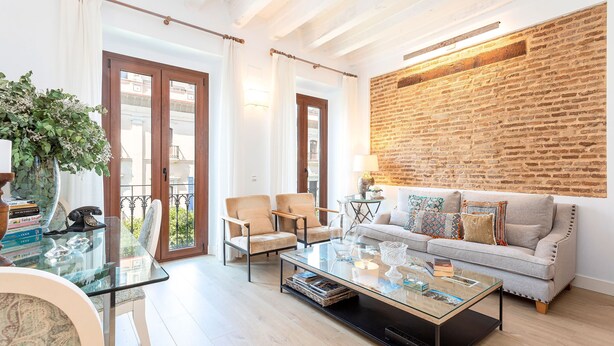 Una sala moderna con una mesa de centro de vidrio, sillones beige, un sofá gris con cojines coloridos y una pared de ladrillo decorativa.