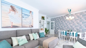 A modern living room with a grey sectional sofa, a glass coffee table, and a dining area with white chairs and a table. - Exclusive Apartment in Front of the sea Victoria (Cádiz)