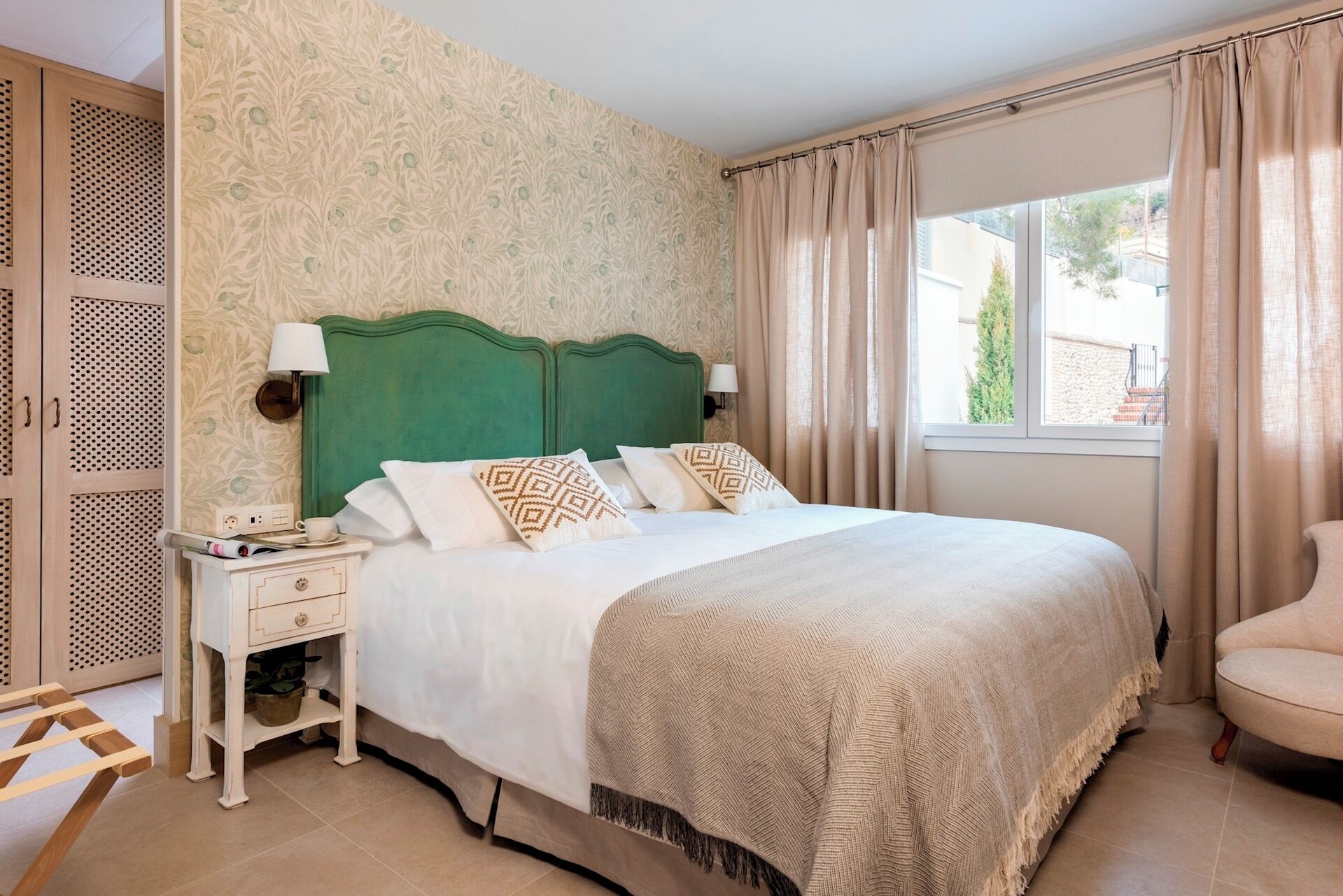 A bedroom with a green headboard, white bedding, a nightstand with a lamp, and a window with curtains.