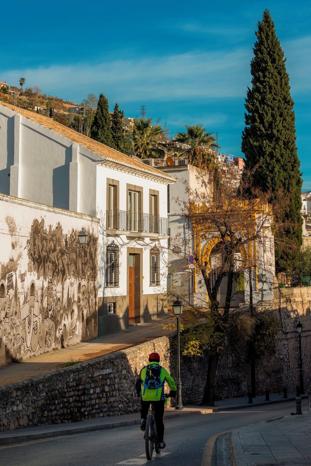 A cyclist on a paved road with a building featuring a balcony and a wall with graffiti.