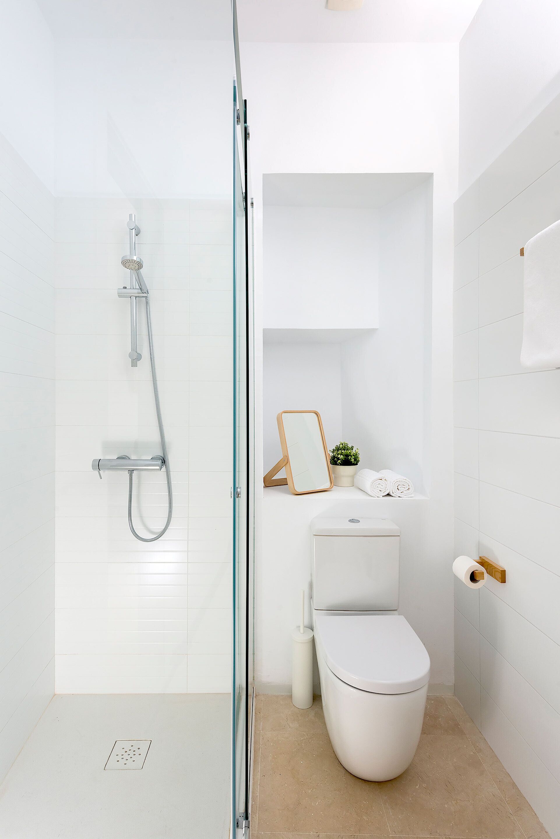 A modern bathroom with a glass-enclosed shower, a toilet, and a small shelf with a mirror, plant, and towels.