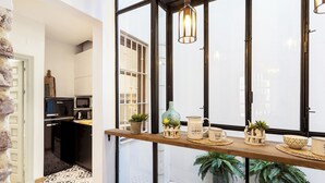 Une cuisine moderne avec une étagère en bois où sont exposés des ustensiles de cuisine et des plantes, un luminaire suspendu et une vue sur la ville à travers de grandes fenêtres.