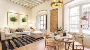 Un salon moderne avec un canapé, un pouf, une table basse et un coin repas avec des chaises. On y trouve un lustre, une plante et une vue sur l’extérieur grâce à de grandes fenêtres.