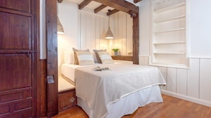 Un dormitorio con puerta de madera, cama con cubrecama blanco, mesita de noche con lámpara y ventana con persiana blanca.